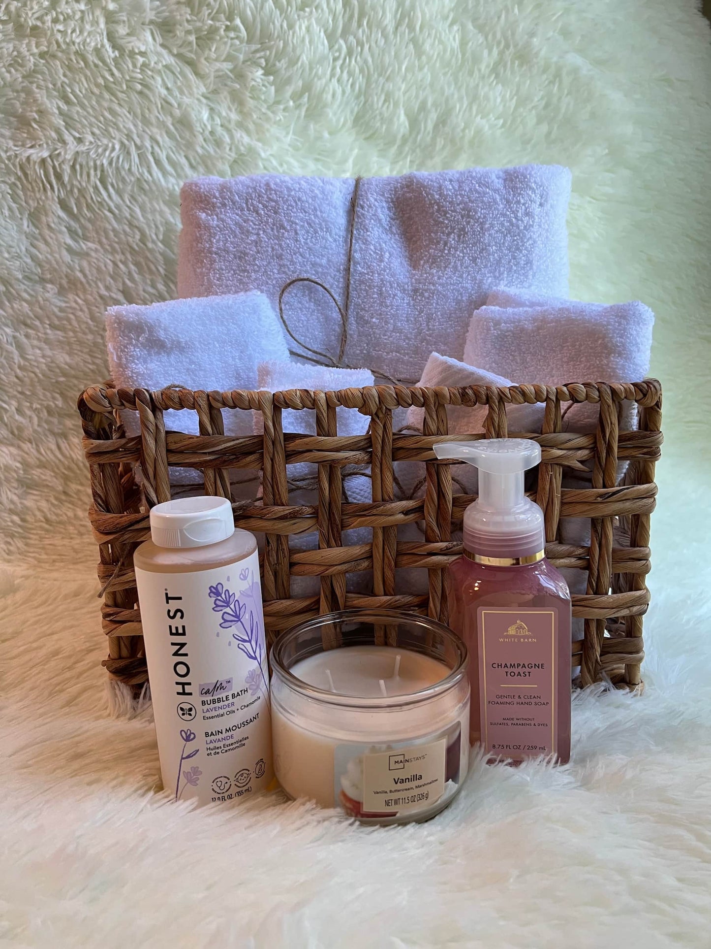 Basket with towels, and personal care products including a candle, hand soap, and a bubble bath bottle on a fluffy white surface.