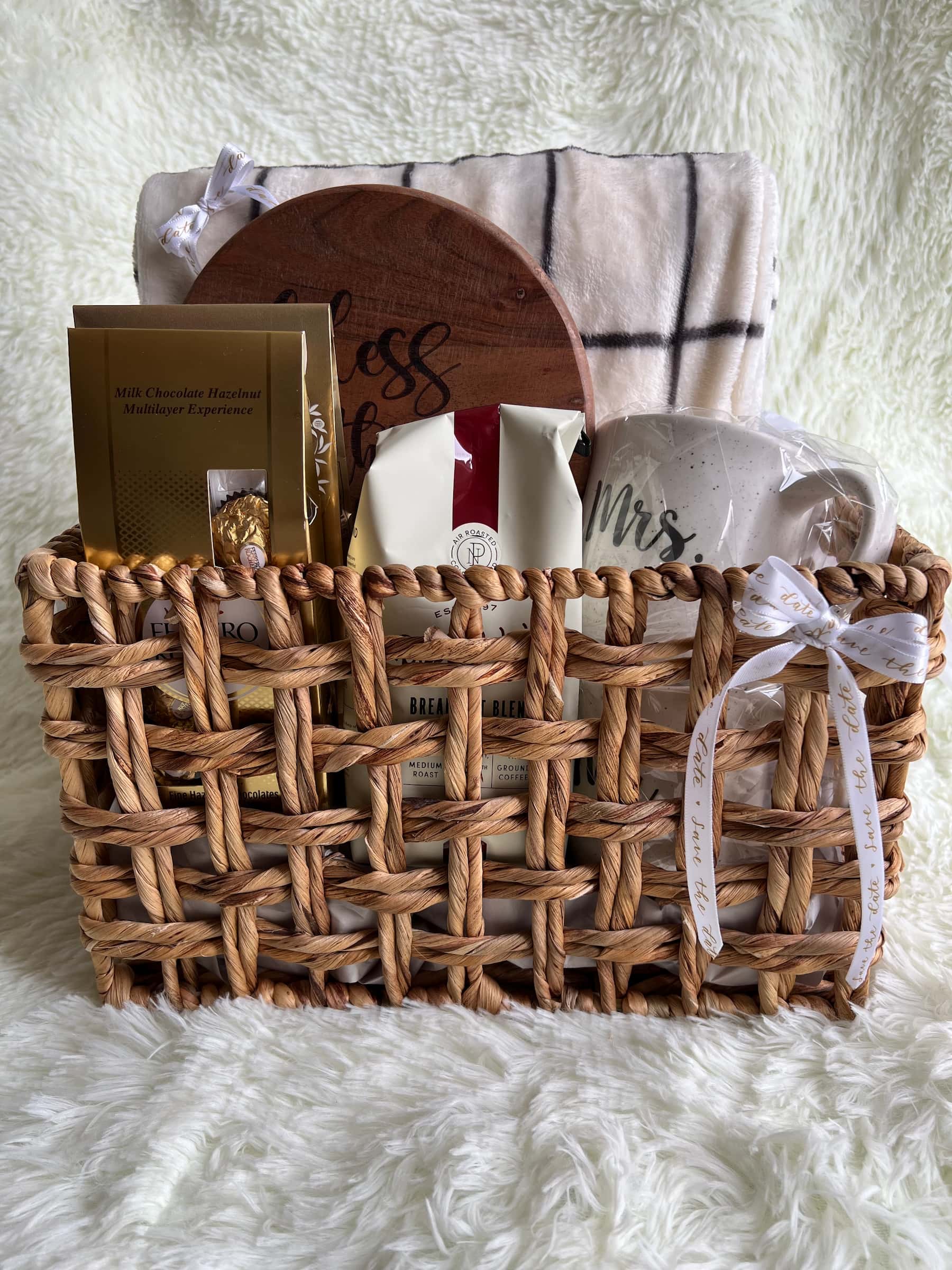 Woven basket with kitchen essentials, including a wooden hot pad, a blanket, chocolate, a matching mug set for newlyweds, and coffee. Decorated with a bow