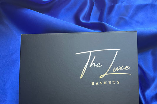 A black square product box with gold foil script reading 'The Luxe Baskets' placed on a shiny royal blue satin fabric background.