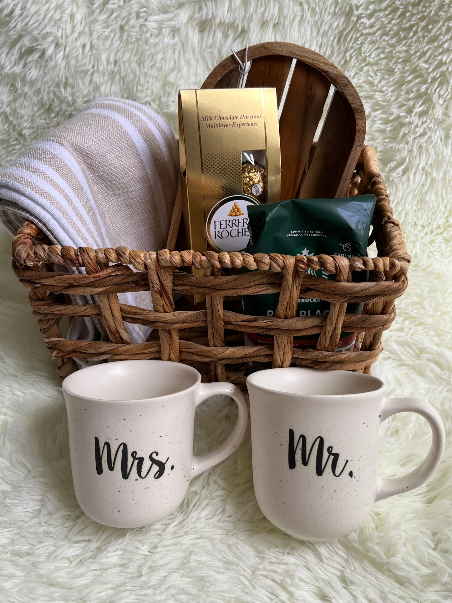Two white mugs labeled 'Mr.' and 'Mrs.' in a wicker basket with chocolate and ground coffee, on a textured surface.
